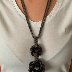 Puede incluir: Un collar de metal gris oscuro con un diseño de doble cadena que conduce a un colgante vertical compuesto por dos nudos de malla texturizada interconectados. La cadena y los nudos comparten un patrón tejido en forma de rejilla constante.