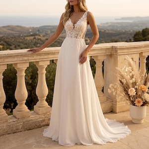 May include: A sleeveless white wedding dress with a V-neckline and a flowing skirt. The bodice features delicate lace detailing. The dress is worn outdoors on a balcony overlooking a scenic landscape. A bouquet of flowers is in a vase.