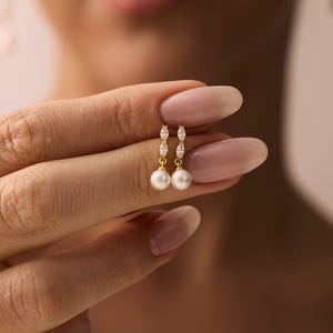 May include: A gold drop earring featuring two small marquise-cut clear stones stacked vertically above a single round white pearl. The earring is held between fingers, showcasing the metallic gold setting and the lustre of the pearl.