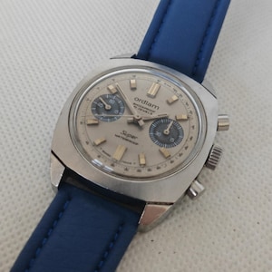 Puede incluir: Un reloj cronógrafo vintage Ordiam con esfera plateada, dos subesferas azul oscuro y caja plateada. El reloj presenta el texto Ordiam, Shockproof, 17 Jewels, Super y Waterproof en la esfera, junto con una correa de cuero de color azul.