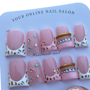 May include: A set of pink press-on nails with a French tip design and leopard print accents. Some nails feature gold rings with colorful rhinestones, while others have small jewel embellishments. The card reads "YOUR ONLINE NAIL SALON."