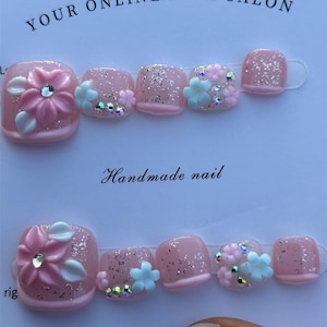 May include: Pink press-on toenails with floral and glitter accents. The nails feature a variety of designs, including 3D flowers, rhinestones, and glitter. The text "YOUR ONLINE NAIL SALON" and "Handmade nail" are visible.