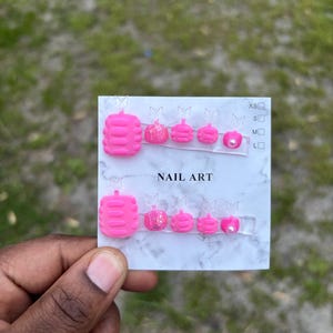 May include: A set of bright pink press-on toenails with various designs, including glitter and a pearl accent. The nails are displayed on a clear card with the text "NAIL ART". Sizes are indicated as XS, S, M, and L.