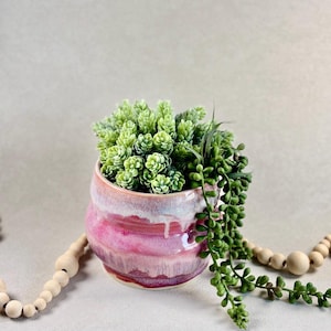 May include: A small, round ceramic planter with a pink and white glaze, filled with green artificial succulents. The planter has a textured, layered design. A strand of faux trailing greenery spills over the side.