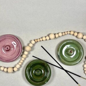 May include: Three ceramic incense holders in shades of pink and green, with a central hole for incense sticks. Two black incense sticks are placed across the holders. A wooden bead garland is also present.