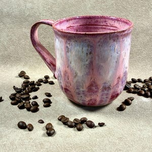 May include: A ceramic mug with a rounded body and a curved handle, featuring a glaze that blends shades of pink, white, and deep magenta in a vertical drip pattern. The mug is surrounded by scattered dark brown roasted coffee beans.