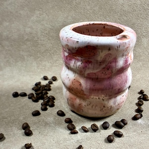 May include: A ceramic cup with a wavy, ribbed silhouette and a speckled glaze featuring blended shades of white, soft pink, and muted terracotta. The cup has a wide, open top and is surrounded by scattered dark brown roasted coffee beans.