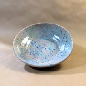 May include: A handmade ceramic bowl with a speckled blue and white glaze. The bowl has a rounded shape and a glossy finish, perfect for serving food or as a decorative piece. The interior and exterior are covered in a speckled pattern.