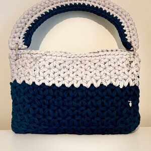Può includere: Una borsa all'uncinetto bicolore con una base blu navy scuro e una sezione superiore grigio chiaro con un motivo a punto a stella. La borsa presenta un manico spesso e arrotondato in colori coordinati e include diversi piccoli ciondoli in metallo argentato.