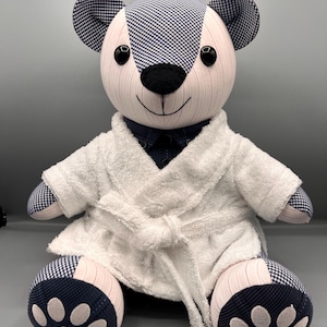 以下が含まれることがあります： 手作りのテディベア。耳、足、頭に白とネイビーのギンガムチェック柄。白いテリークロスのバスローブとネイビーのシャツを着ています。クマの顔は薄いピンク色で、黒い鼻とボタンの目があります。