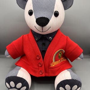 May include: A plush teddy bear dressed in a red blazer with three buttons, a dark blue shirt, and a black and white gingham pattern on the ears and paws. The blazer features a "Deighton Gates" patch.