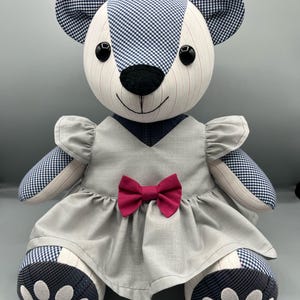 Peut inclure: Un ours en peluche composé d'un patchwork de tissu à carreaux vichy bleu et à rayures blanches. L'ours porte une robe grise à manches volantées avec un petit nœud magenta au centre. Il possède des yeux en boutons noirs et un nez noir. Texte : Memories Made by Me.