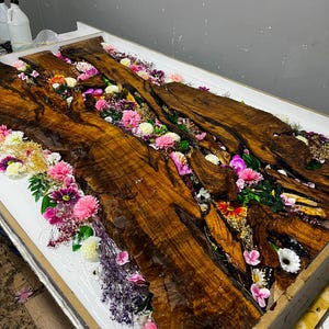 May include: A rectangular wooden table top featuring two live-edge walnut slabs with a central river-like channel filled with clear resin, embedded with a variety of colorful artificial flowers including pink, white, and purple blossoms.