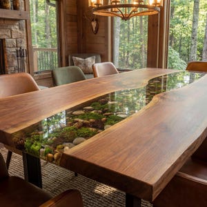 May include: A rectangular dining table featuring two thick live-edge walnut wood slabs separated by a central transparent resin inlay. The resin section contains embedded green moss, small stones, and pinecones. The table stands on dark metal legs.