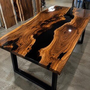 Peut inclure: Une table à manger rectangulaire composée d'une dalle de bois à bord naturel avec une rivière centrale en résine époxy noire brillante. La table est soutenue par une base en métal noir avec des pieds rectangulaires et une finition polie.