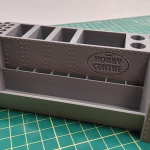 May include: A grey plastic hobby organiser with various compartments. It has sections for paintbrushes, rectangular slots, and cylindrical holes. The text "THE HOBBY CENTRE" is embossed on the side. Ideal for storing hobby supplies.