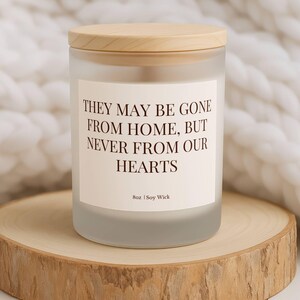 Puede incluir: Una vela de vidrio esmerilado con tapa de madera, con la inscripci&oacute;n "THEY MAY BE GONE FROM HOME, BUT NEVER FROM OUR HEARTS". La vela est&aacute; sobre una rodaja de madera, con una manta blanca tejida en el fondo. La vela es de 227g.