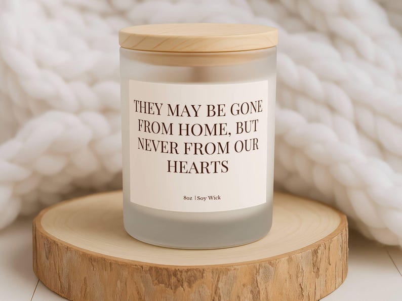 Puede incluir: Una vela de vidrio esmerilado con tapa de madera, con la inscripci&oacute;n "THEY MAY BE GONE FROM HOME, BUT NEVER FROM OUR HEARTS". La vela est&aacute; sobre una rodaja de madera, con una manta blanca tejida en el fondo. La vela es de 227g.