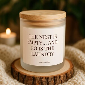 Puede incluir: Un tarro de vela de vidrio esmerilado con tapa de madera, con el texto "THE NEST IS EMPTY... AND SO IS THE LAUNDRY" en una etiqueta blanca. La vela est&aacute; sobre un posavasos de madera, con una vela encendida y una manta de punto en el fondo.