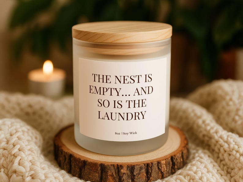 Puede incluir: Un tarro de vela de vidrio esmerilado con tapa de madera, con el texto "THE NEST IS EMPTY... AND SO IS THE LAUNDRY" en una etiqueta blanca. La vela est&aacute; sobre un posavasos de madera, con una vela encendida y una manta de punto en el fondo.