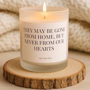 Puede incluir: Una vela encendida en un tarro de vidrio esmerilado con el texto "THEY MAY BE GONE FROM HOME, BUT NEVER FROM OUR HEARTS." La vela est&aacute; sobre una rodaja de madera. La vela es de 227g y est&aacute; hecha con mecha de soja.
