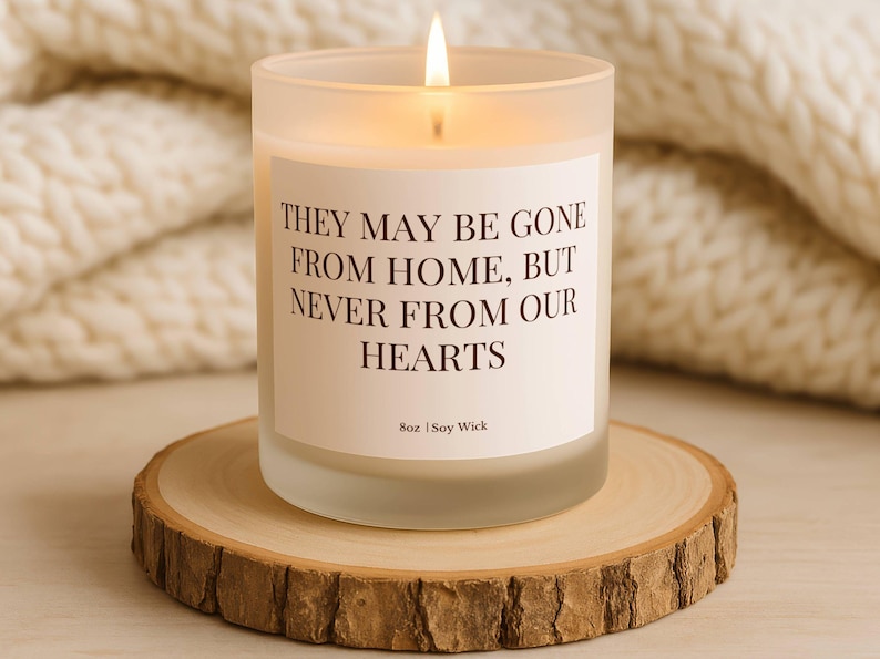 Puede incluir: Una vela encendida en un tarro de vidrio esmerilado con el texto "THEY MAY BE GONE FROM HOME, BUT NEVER FROM OUR HEARTS." La vela est&aacute; sobre una rodaja de madera. La vela es de 227g y est&aacute; hecha con mecha de soja.
