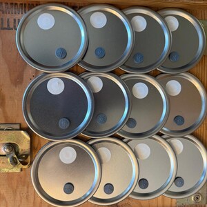 May include: A collection of silver-colored metal lids with a white circular patch and a small gray fastener. The lids are arranged in rows on a wooden surface, suggesting a storage or organizational context.