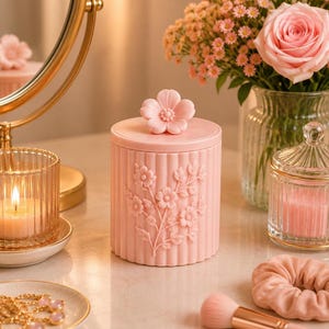May include: A cylindrical light pink storage jar with a vertical ribbed texture and a raised floral relief pattern on the front. The lid features a sculpted flower handle. The container is made of a solid material and has a smooth matte finish.