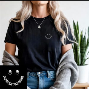 Puede incluir: Camiseta negra con un pequeño diseño de carita sonriente que dice "Pharmacy Technician" en una fuente curva. El diseño está en la parte delantera de la camiseta. Una versión más grande del diseño está en la esquina inferior izquierda de la imagen.