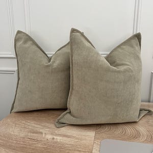 May include: Two beige cushions with a herringbone pattern. The cushions are made of a soft fabric and are placed on a wooden surface. The cushions have a classic, neutral design.