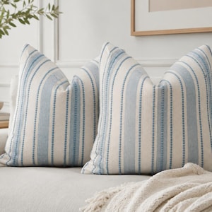 Peut inclure: Deux coussins carrés décoratifs avec un motif à rayures verticales bleu clair et blanc cassé. Le tissu présente une alternance de larges bandes bleu clair et de fines lignes texturées en pointillés bleus sur un fond blanc cassé.