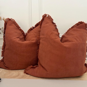 May include: Two terracotta-coloured cushions with a herringbone pattern and fringed edges. The cushions are made of a soft, textured fabric and are arranged side by side. The cushions are a warm, inviting accent for any living space.