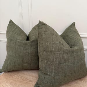 May include: Two square throw cushions in a muted olive green colour. The fabric features a visible woven texture with a subtle grid pattern created by stitched lines across the surface. The cushions have a structured, plump appearance with clean edges.