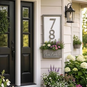 May include: A rectangular vertical house number sign with a gray frame and white background displaying the number 78 in large gray digits. Below the numbers is a built-in planter box filled with colorful flowers and the text Arce Acres Ln.