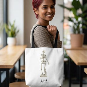 Può includere: Borsa tote bianca con un'illustrazione dettagliata di uno scheletro umano. Lo scheletro è in piedi su un piedistallo. La parola "Hal" è stampata in nero sotto l'illustrazione. La borsa ha cinghie nere.