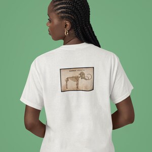 May include: White t-shirt featuring a vintage-style illustration of an elephant skeleton. The image is in a rectangular frame on the back of the shirt. The shirt is plain and has short sleeves. The background is a solid green color.