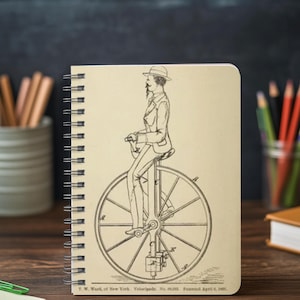 May include: Spiral-bound notebook featuring a vintage illustration of a man riding a high-wheel bicycle. The black and white drawing includes the text "T.W. Ward, of New York. Velocipede. No. 88,003. Patented April 6, 1869."