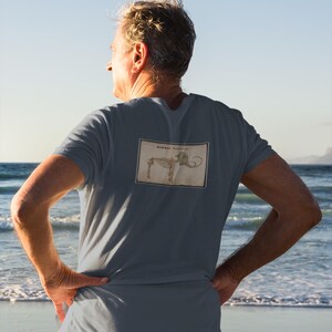 May include: A slate blue t-shirt featuring a vintage illustration of a mammoth skeleton. The rectangular graphic is centered on the back of the shirt. The person in the image is standing on a beach with the ocean in the background.