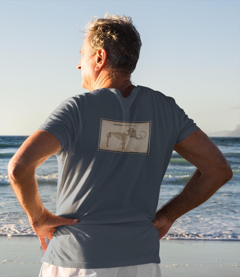 May include: A slate blue t-shirt featuring a vintage illustration of a mammoth skeleton. The rectangular graphic is centered on the back of the shirt. The person in the image is standing on a beach with the ocean in the background.