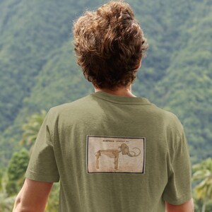 May include: Olive green t-shirt featuring a vintage illustration of an elephant skeleton within a rectangular frame. The text "ELEPHAS" is visible above the illustration. The shirt is made of a soft, comfortable material.