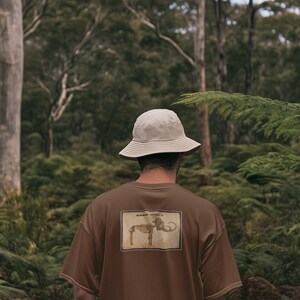 May include: A brown t-shirt with a vintage-style illustration of an elephant skeleton on the back. The person is wearing a beige bucket hat. The background is a lush green forest.