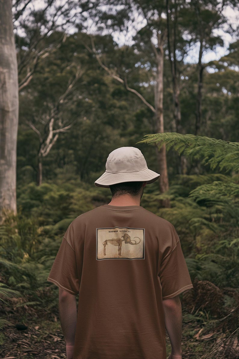 May include: A brown t-shirt with a vintage-style illustration of an elephant skeleton on the back. The person is wearing a beige bucket hat. The background is a lush green forest.