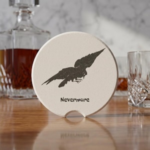 May include: A round, off-white stone coaster featuring a black silhouette of a flying raven and the text "Nevermore" printed in a dark, sans-serif font. The coaster has a small, semi-circular notch cut out from the bottom edge of its circular frame.