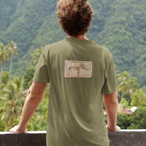 May include: Olive green t-shirt with a rectangular graphic of a mammoth skeleton on the back. The graphic is in a vintage style with brown and beige tones. The person is wearing blue jeans and is standing outside.