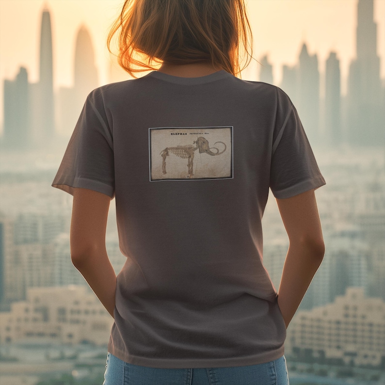 May include: Gray t-shirt with a rectangular graphic of an elephant skeleton labeled "ELEPHAS antiquus" on the back. The shirt is a muted gray color and has short sleeves. The background shows a cityscape at sunset.