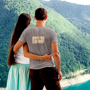 May include: A gray t-shirt with a rectangular graphic on the back, worn by a person. The person is embracing another person, with a scenic mountain and lake backdrop. The t-shirt graphic appears to be a vintage illustration.