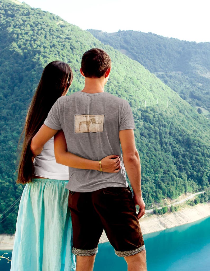 May include: A gray t-shirt with a rectangular graphic on the back, worn by a person. The person is embracing another person, with a scenic mountain and lake backdrop. The t-shirt graphic appears to be a vintage illustration.