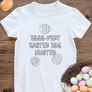 May include: White t-shirt with the text "EGGS-PERT EASTER EGG HUNTER" in black. The shirt features three egg designs. A decorative nest with colorful Easter eggs is in the lower right corner. An orange egg is in the upper left corner.