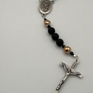 Puede incluir: Un rosario con cuentas de piedra de lava negra, cuentas doradas y un crucifijo plateado. La pieza central representa un ángel de la guarda. Este artículo religioso se utiliza para la oración y la meditación.