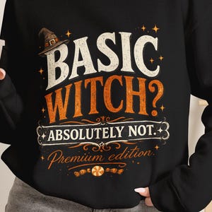 May include: A black long-sleeve sweatshirt featuring a graphic print with the text "BASIC WITCH? ABSOLUTELY NOT. Premium edition" in white and orange. The design includes a small witch hat, decorative flourishes, and stars on the front chest.